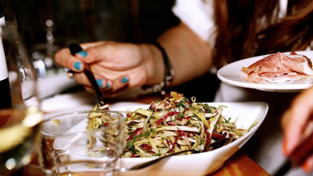 Köstlicher Salat welcher auf einem Wohnmobil Tisch steht und aus dem von einer weiblichen Hand gerade eine Gabel voll Salat auf einen Teller mit Serano Schinken gehoben wird.